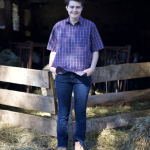 Mabe (they/them) on their farm standing in front of a pen with a wooden gate and sheep are inside the pen. Mabe is wearing a purple plaid button up short sleeved shirt, jeans, and work boots.